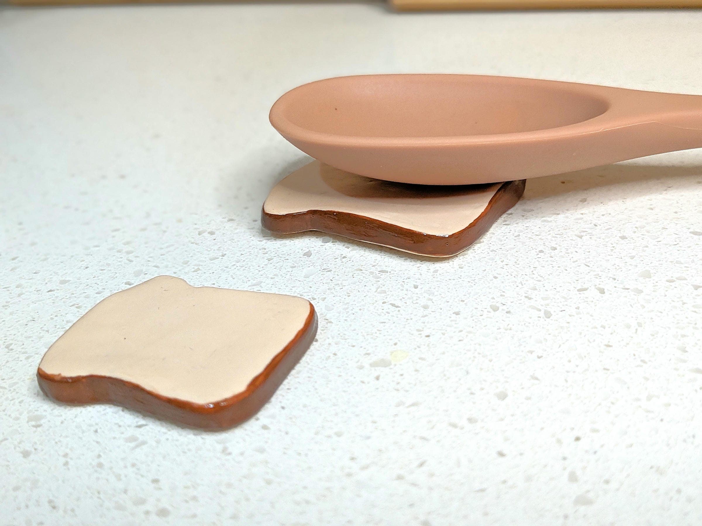 A ceramic spoon rest shaped like toast, with one piece of toast under the spoon and another beside it, on a speckled countertop.