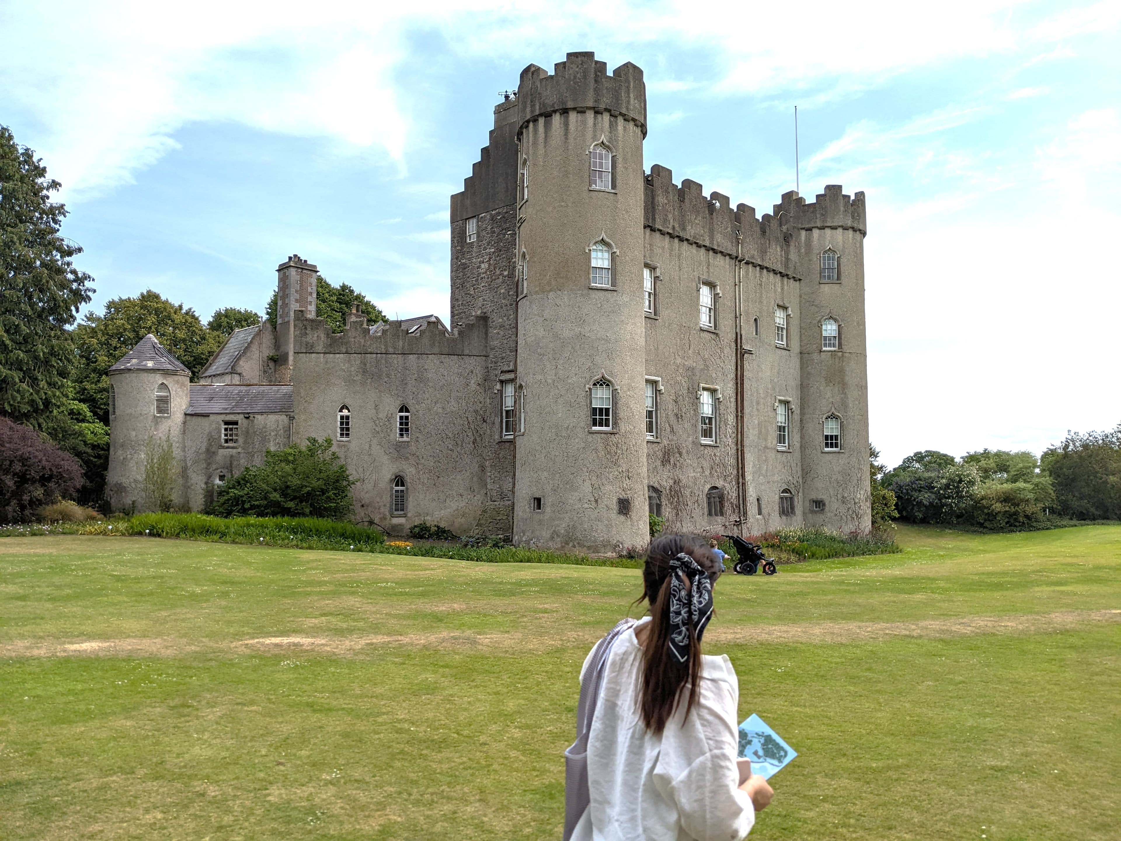 Person holding a brochure, standing on a lawn in front of a historic stone castle with round towers and multiple windows.