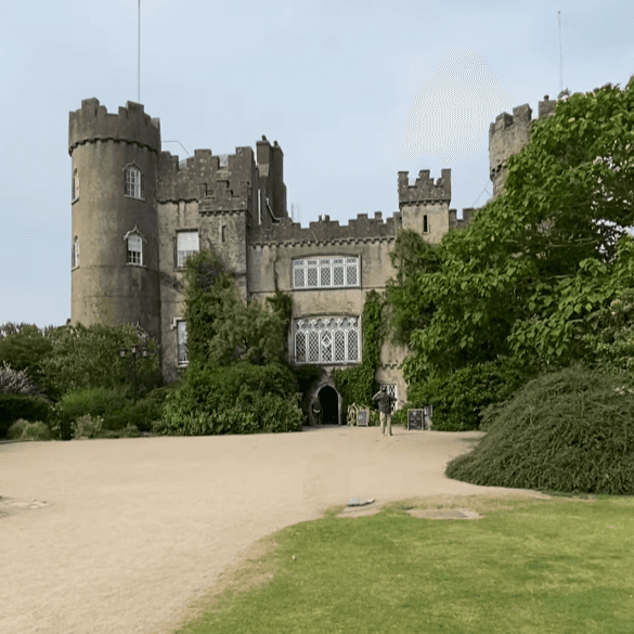 Historic stone castle with round turrets and ivy-covered walls, set against a clear sky, surrounded by lush gardens and a gravel path.