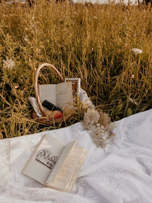 Open books on a white lace cloth in a grassy field with a wicker basket and dried flowers.