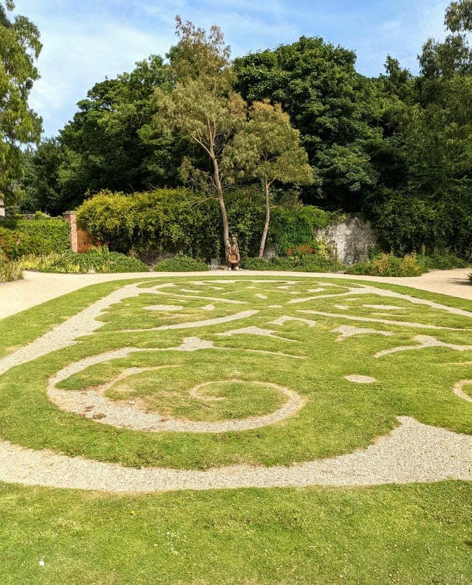 Intricate garden with swirling grass patterns and surrounding trees under a clear blue sky.