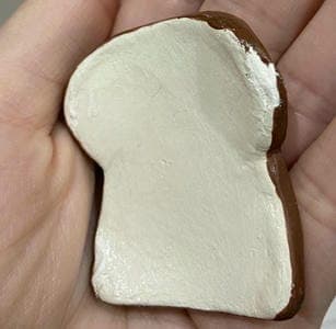 A person holding a small, realistic slice of bread sculpture with a white top and brown crust edges.
